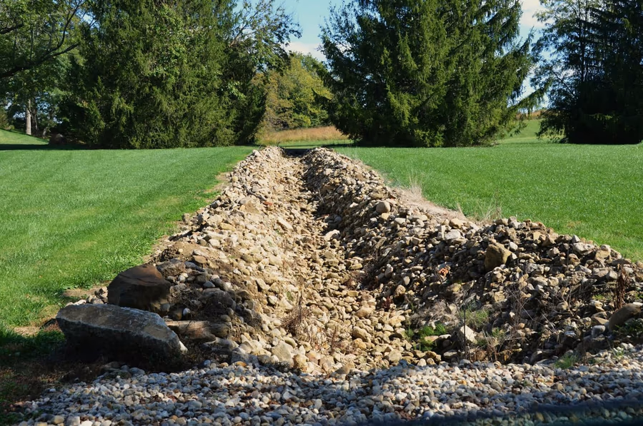 A rock-lined drainage channel cut across a mown lawn, running between stands of cedar trees